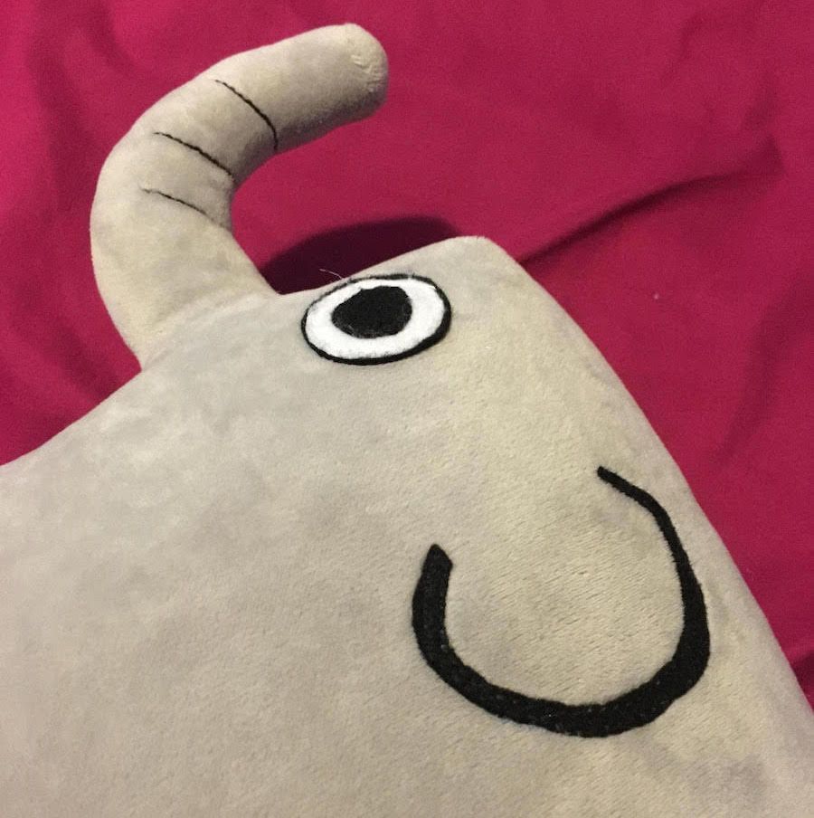 Select to view 'Close-up of Leo the Elephant plush pillow's face, highlighting detailed embroidered eye, smiling mouth, and curved stuffed trunk on soft grey fabric.' image
