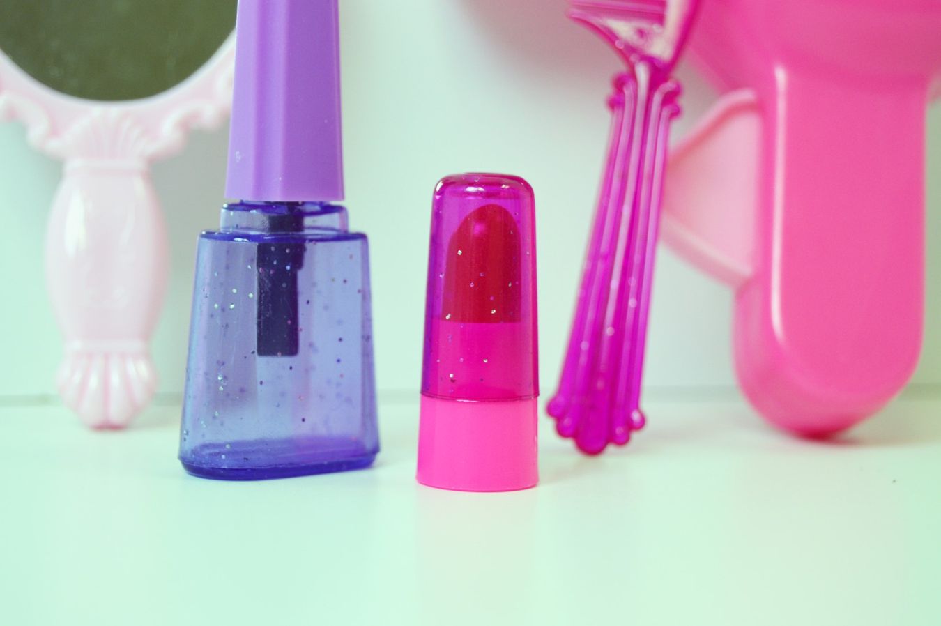 Select to view 'Close-up of purple and pink toy nail polish bottles with a pink plastic comb and decorative pink handheld mirror in soft background lighting.' image