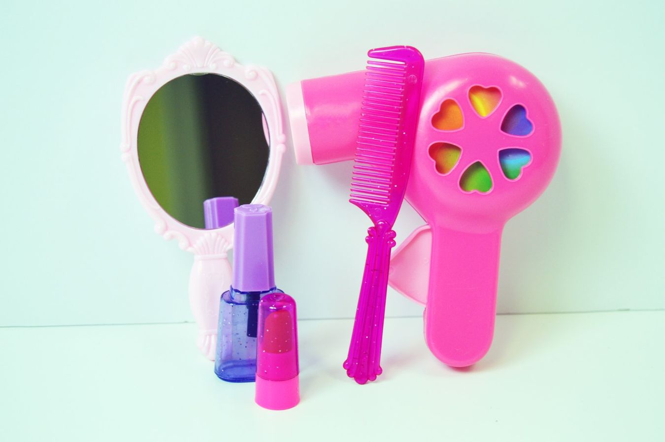 Select to view 'Bright kids' beauty set featuring pink toy hairdryer with heart-shaped openings, matching plastic comb, toy nail polish bottles, and an ornate pink framed mirror.' image